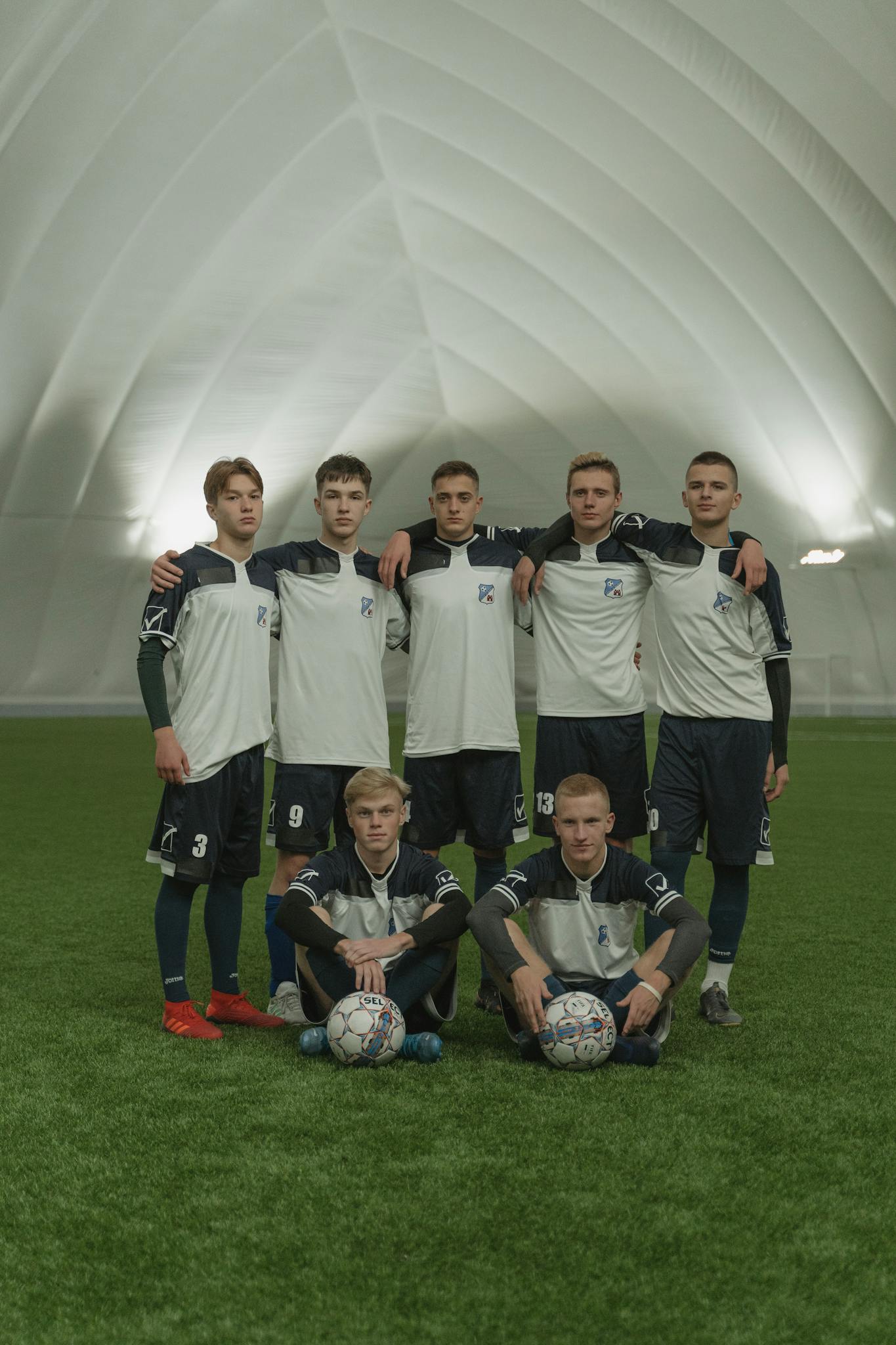 A group of young male soccer players posing indoors on a sports field with two soccer balls.