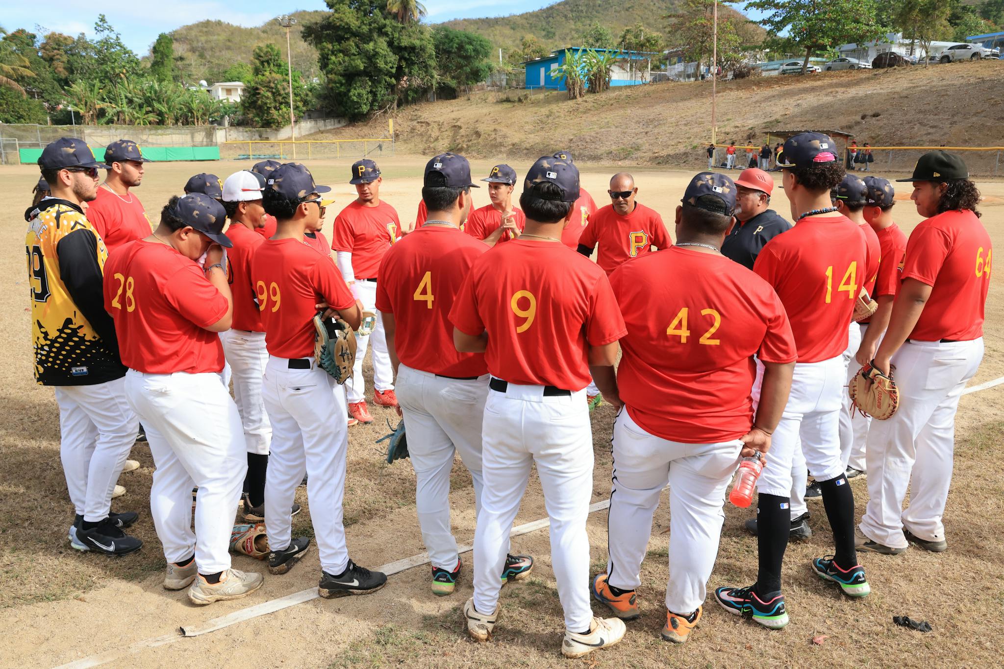 Baseball team in red jerseys gathers for a pre-game strategy session on a sunny day.